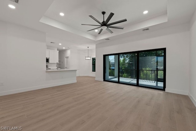 unfurnished living room with ceiling fan with notable chandelier, light wood-type flooring, and a raised ceiling