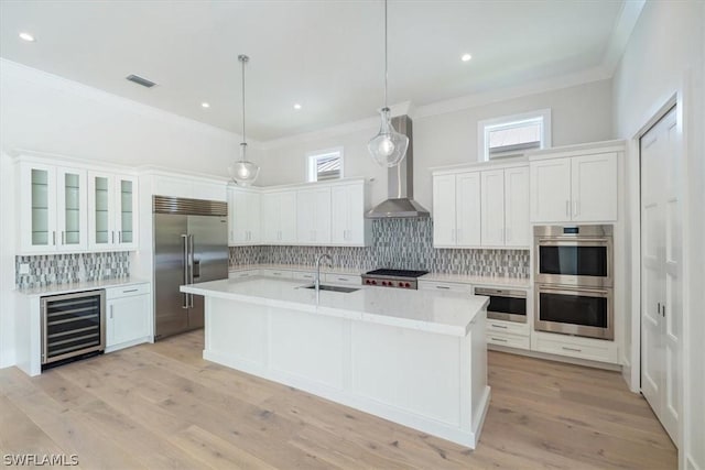 kitchen featuring hanging light fixtures, wall chimney exhaust hood, an island with sink, appliances with stainless steel finishes, and beverage cooler