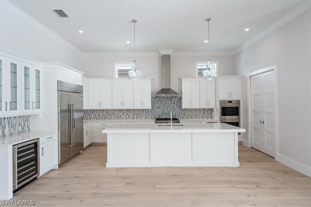 kitchen featuring light wood-type flooring, stainless steel appliances, beverage cooler, wall chimney range hood, and hanging light fixtures