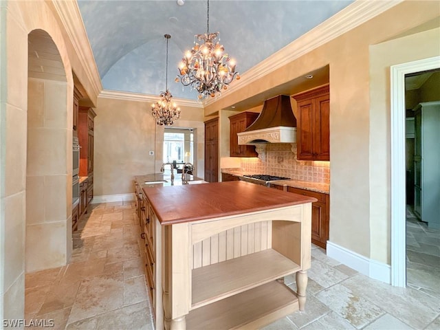 kitchen with custom exhaust hood, a center island with sink, crown molding, hanging light fixtures, and a chandelier