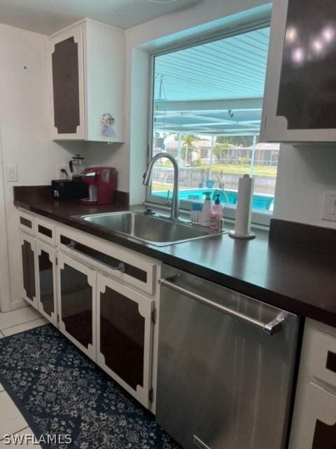 kitchen featuring dark countertops, light tile patterned floors, stainless steel dishwasher, white cabinets, and a sink
