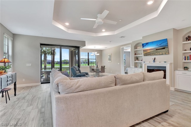 living room with a raised ceiling, built in features, light hardwood / wood-style flooring, and ceiling fan with notable chandelier