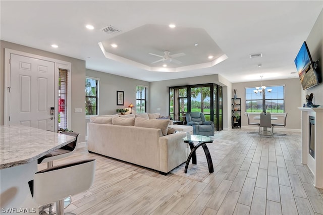 living room with a raised ceiling, a healthy amount of sunlight, ceiling fan with notable chandelier, and light wood-type flooring