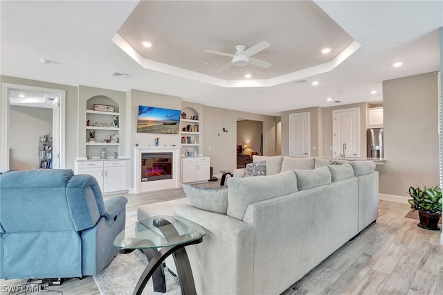 living room with built in shelves, a raised ceiling, ceiling fan, sink, and light hardwood / wood-style flooring