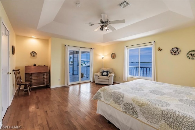 bedroom with ceiling fan, a raised ceiling, dark wood-type flooring, access to exterior, and a water view