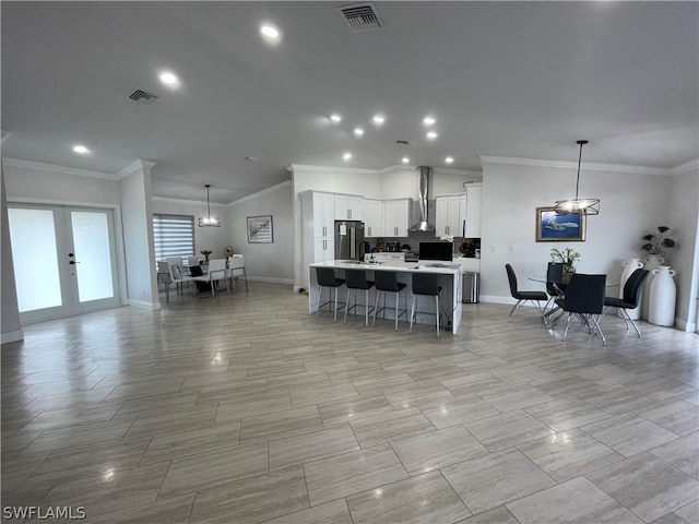 kitchen with wall chimney range hood, stainless steel refrigerator, white cabinetry, hanging light fixtures, and an island with sink