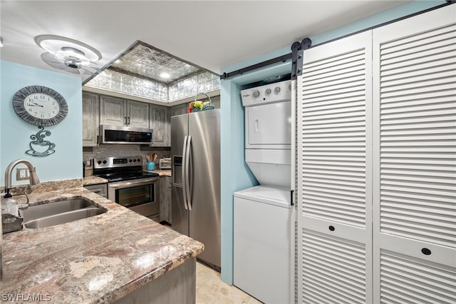 kitchen featuring stainless steel appliances, a sink, backsplash, light stone countertops, and stacked washer and clothes dryer