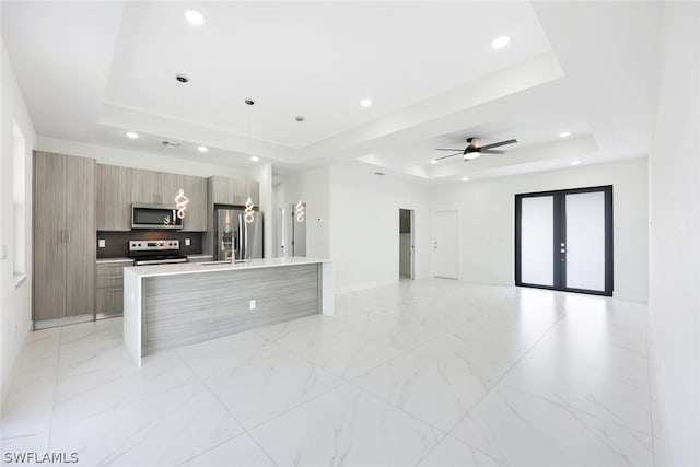 kitchen with ceiling fan, hanging light fixtures, a raised ceiling, a center island with sink, and appliances with stainless steel finishes