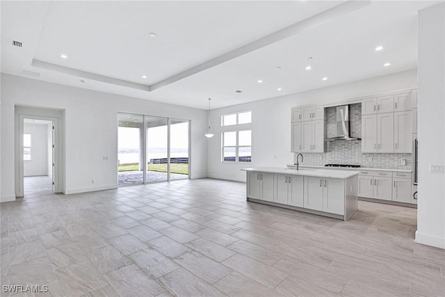 kitchen with tasteful backsplash, sink, white cabinets, wall chimney range hood, and a center island with sink