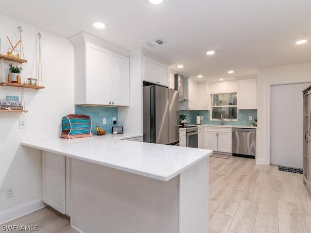 kitchen with tasteful backsplash, white cabinets, kitchen peninsula, stainless steel appliances, and wall chimney exhaust hood