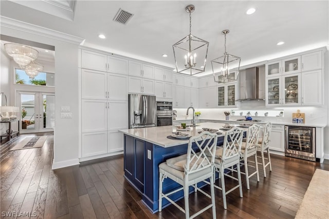 kitchen with wine cooler, light countertops, wall chimney range hood, appliances with stainless steel finishes, and glass insert cabinets