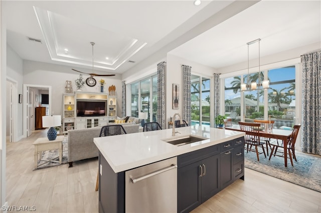 kitchen with light hardwood / wood-style floors, a raised ceiling, an island with sink, stainless steel dishwasher, and sink