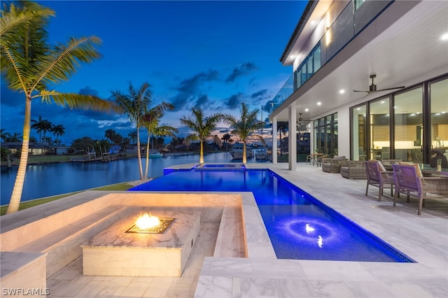 pool at dusk with a water view, ceiling fan, an in ground hot tub, an outdoor living space with a fire pit, and a patio area