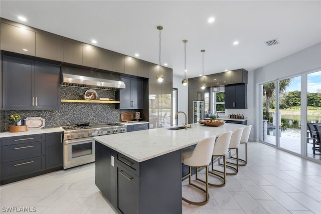 kitchen with decorative backsplash, ventilation hood, a kitchen island with sink, decorative light fixtures, and double oven range