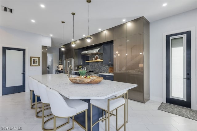 kitchen featuring light stone countertops, tasteful backsplash, pendant lighting, a kitchen island with sink, and exhaust hood