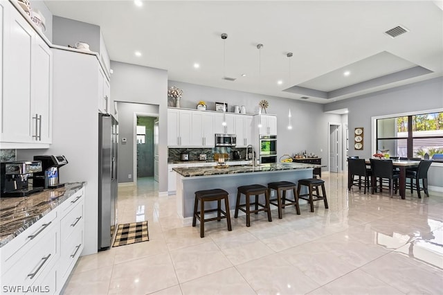 kitchen with a kitchen island with sink, dark stone counters, a raised ceiling, white cabinetry, and stainless steel appliances