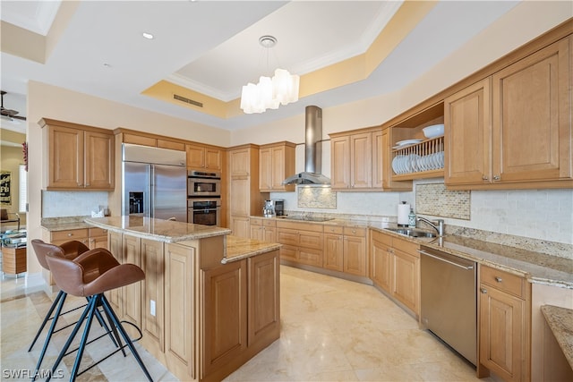 kitchen featuring pendant lighting, stainless steel appliances, a tray ceiling, a kitchen island, and wall chimney exhaust hood