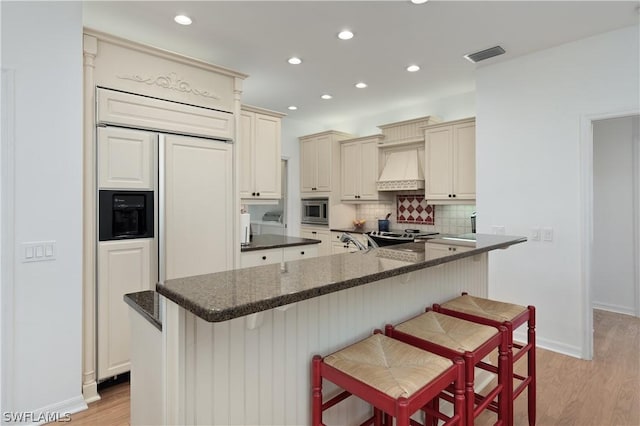 kitchen featuring cream cabinetry, custom range hood, built in appliances, and a breakfast bar