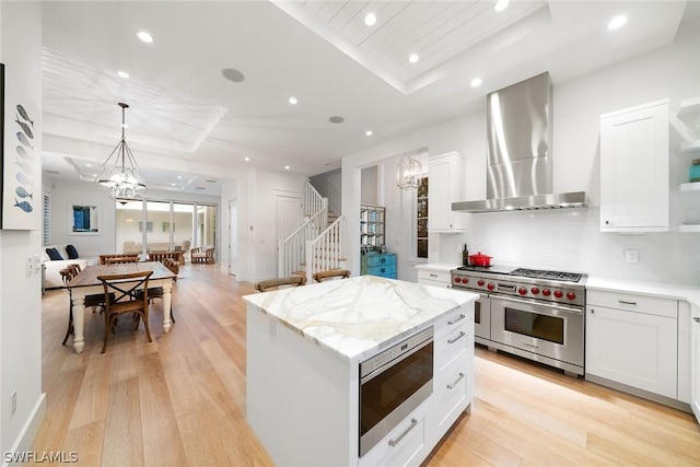 kitchen with wall chimney exhaust hood, stainless steel appliances, a center island, white cabinetry, and an inviting chandelier