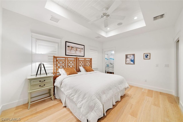 bedroom with a tray ceiling, ceiling fan, light hardwood / wood-style floors, and connected bathroom