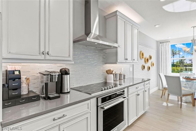 kitchen featuring stainless steel oven, wall chimney exhaust hood, hanging light fixtures, black electric cooktop, and light wood-type flooring