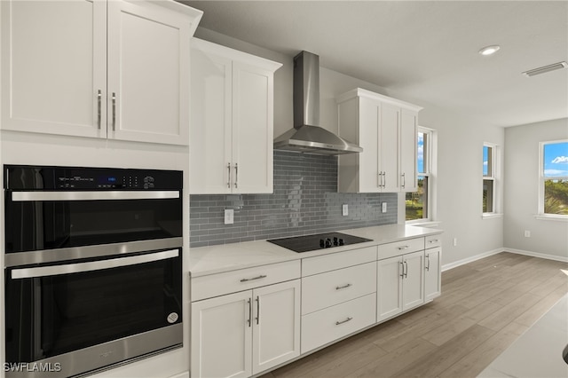 kitchen featuring backsplash, multiple ovens, wall chimney range hood, light countertops, and black electric cooktop