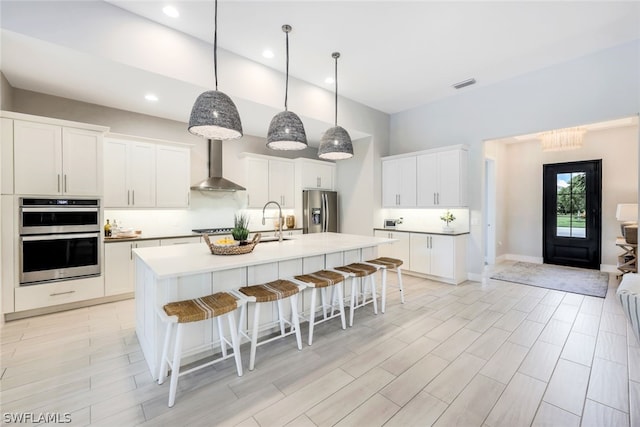 kitchen with white cabinets, appliances with stainless steel finishes, a kitchen island with sink, and wall chimney range hood