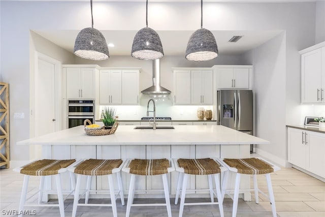 kitchen featuring wall chimney exhaust hood, stainless steel appliances, hanging light fixtures, an island with sink, and white cabinets