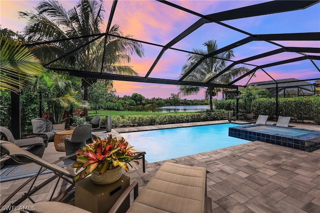 pool at dusk with glass enclosure, an in ground hot tub, a patio area, and a water view