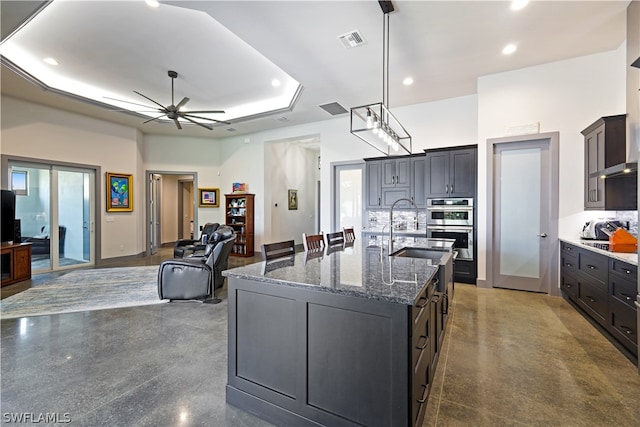 kitchen with dark stone counters, ceiling fan, a raised ceiling, a kitchen island with sink, and sink