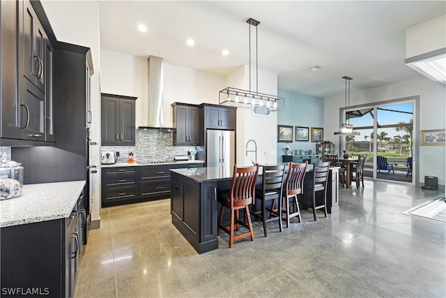 kitchen with decorative light fixtures, stainless steel refrigerator, light stone counters, backsplash, and an island with sink