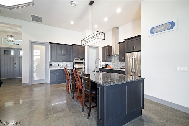 kitchen featuring dark brown cabinets, tasteful backsplash, stainless steel appliances, dark stone countertops, and a kitchen island with sink