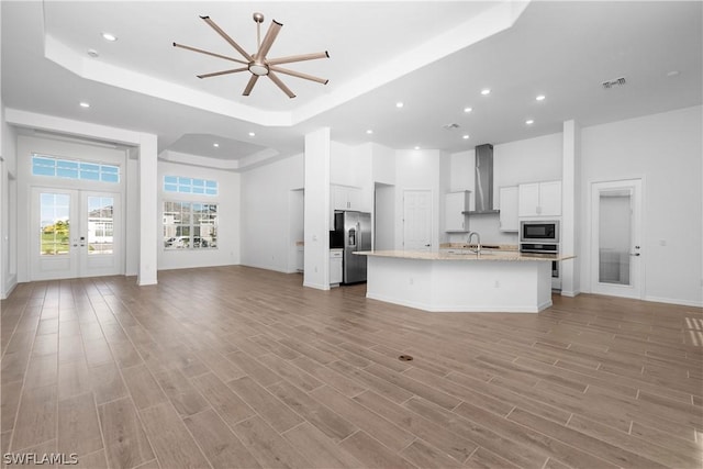 kitchen featuring white cabinets, appliances with stainless steel finishes, wall chimney range hood, an island with sink, and a tray ceiling