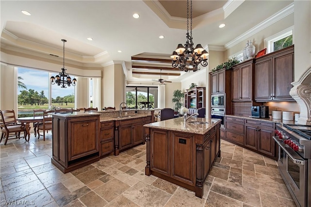 kitchen featuring ceiling fan with notable chandelier, stainless steel appliances, a raised ceiling, pendant lighting, and an island with sink