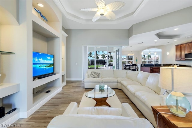 living room featuring a tray ceiling, ceiling fan, crown molding, built in features, and light hardwood / wood-style flooring