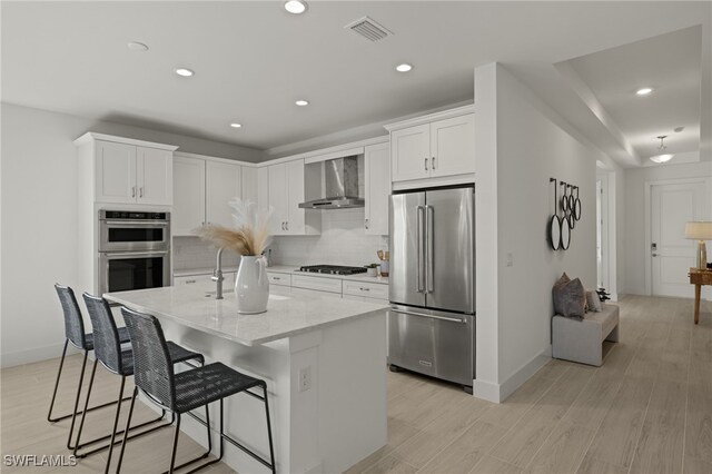 kitchen with appliances with stainless steel finishes, white cabinetry, an island with sink, light stone countertops, and wall chimney range hood