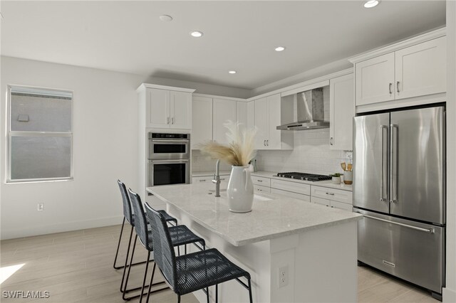 kitchen featuring wall chimney exhaust hood, stainless steel appliances, an island with sink, and white cabinets