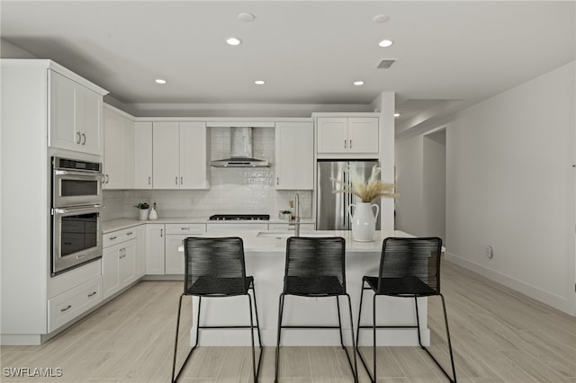 kitchen with sink, white cabinets, a kitchen island with sink, stainless steel appliances, and wall chimney range hood