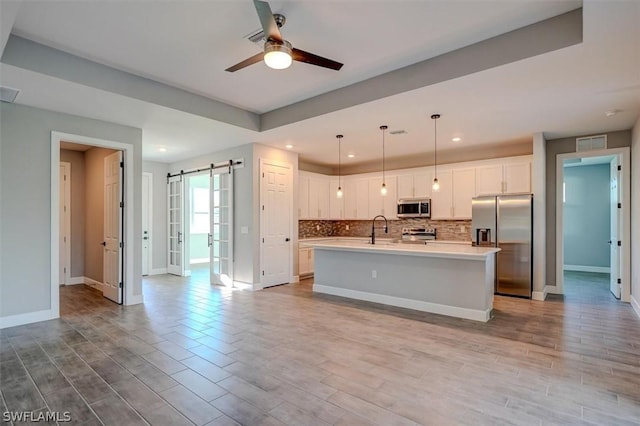 kitchen with hanging light fixtures, stainless steel appliances, a barn door, a kitchen island with sink, and white cabinets