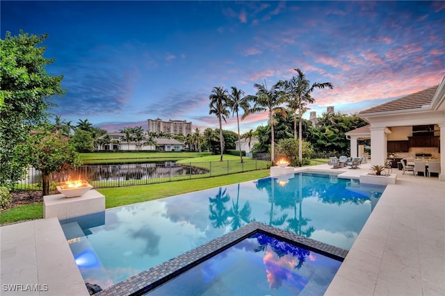 pool at dusk featuring a water view, a patio, and a lawn