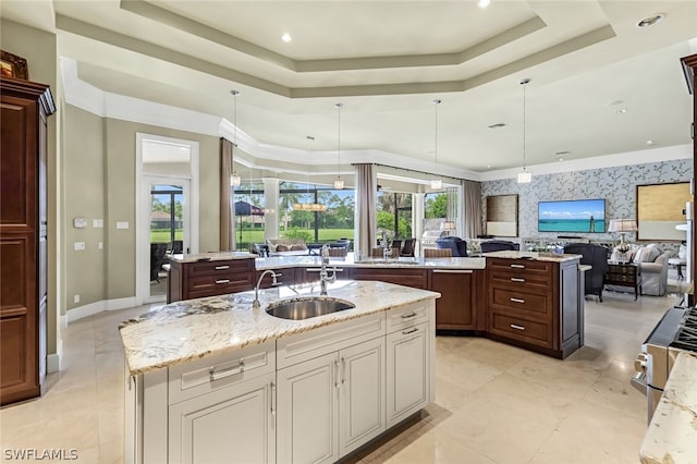 kitchen featuring sink, a tray ceiling, light stone countertops, an island with sink, and decorative light fixtures