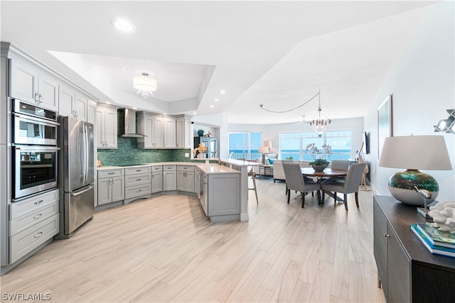 kitchen with wall chimney range hood, pendant lighting, light wood-type flooring, a raised ceiling, and appliances with stainless steel finishes