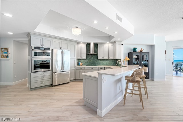 kitchen with light wood-type flooring, kitchen peninsula, stainless steel appliances, wall chimney exhaust hood, and a kitchen breakfast bar