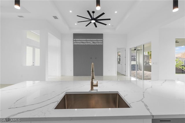 kitchen with a raised ceiling, ceiling fan, light stone counters, and sink