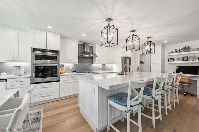 kitchen with stainless steel appliances, wall chimney range hood, a center island with sink, white cabinets, and hanging light fixtures