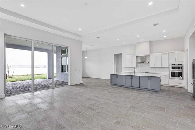 kitchen featuring wall chimney exhaust hood, double oven, white cabinets, a kitchen island with sink, and backsplash