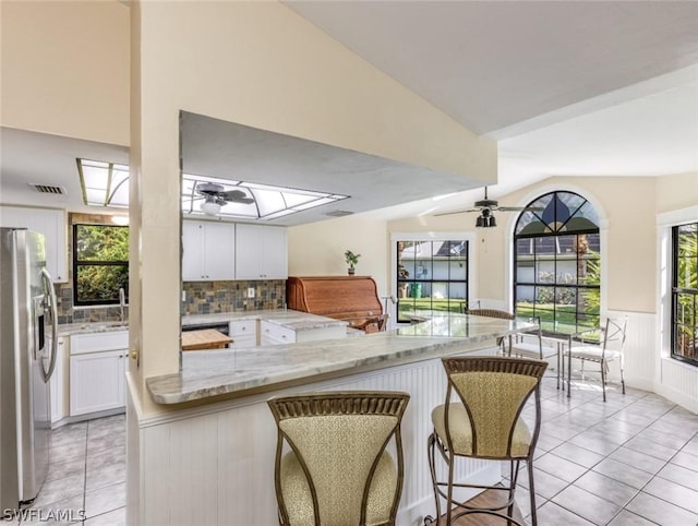 kitchen with kitchen peninsula, stainless steel fridge with ice dispenser, light stone counters, a kitchen bar, and white cabinetry