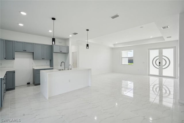 kitchen with sink, gray cabinetry, hanging light fixtures, a center island with sink, and a raised ceiling