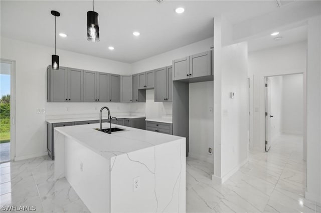 kitchen featuring sink, gray cabinetry, light stone counters, decorative light fixtures, and a center island with sink