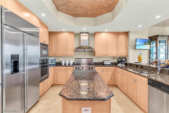 kitchen with sink, appliances with stainless steel finishes, a kitchen island, a raised ceiling, and wall chimney exhaust hood